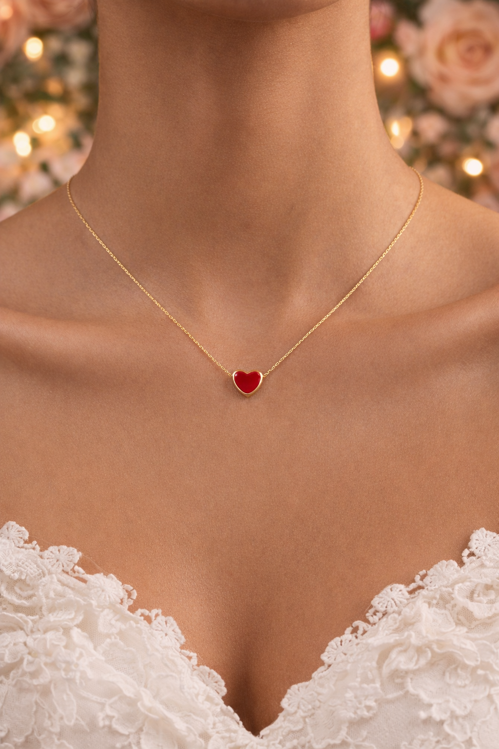 Gold necklace with a red heart pendant on a person wearing a white lace garment, with a blurred floral and light background.